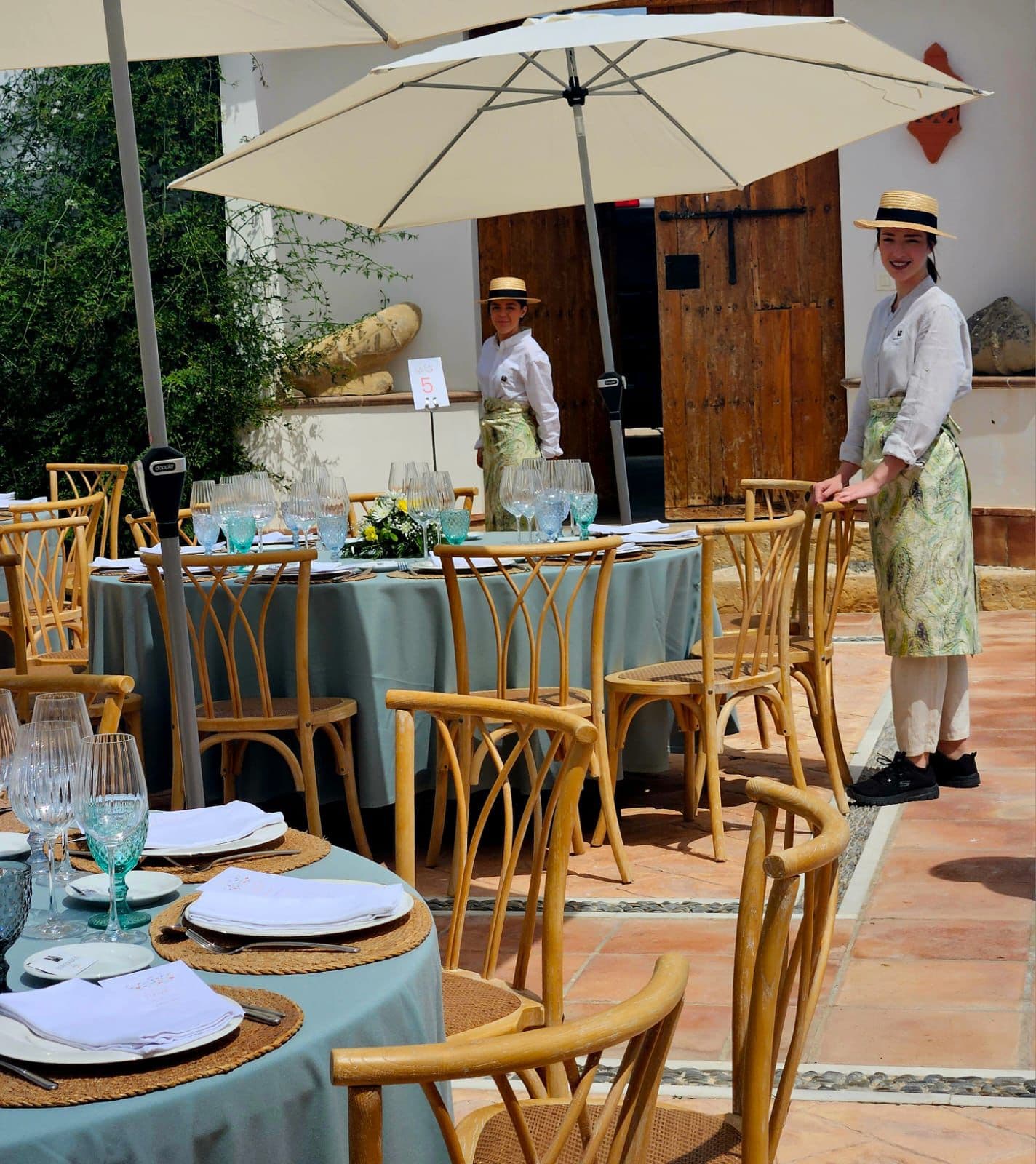 Mesa alta de aperitivo con mantel verde agua bajo una sombrilla, con camareros de canotier atendiendo