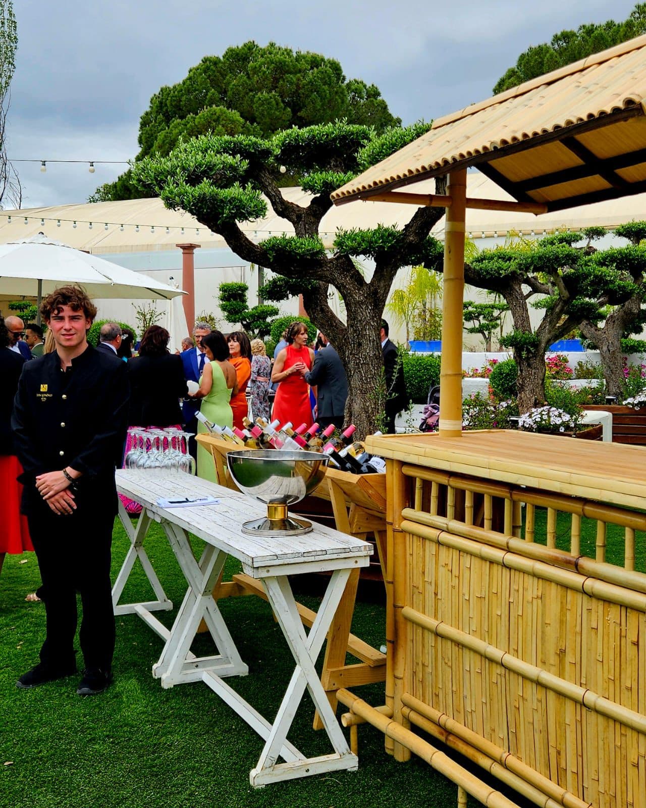 Mesa de dulces junto al bonsái del jardín, bajo una pérgola de madera, con invitados alrededor