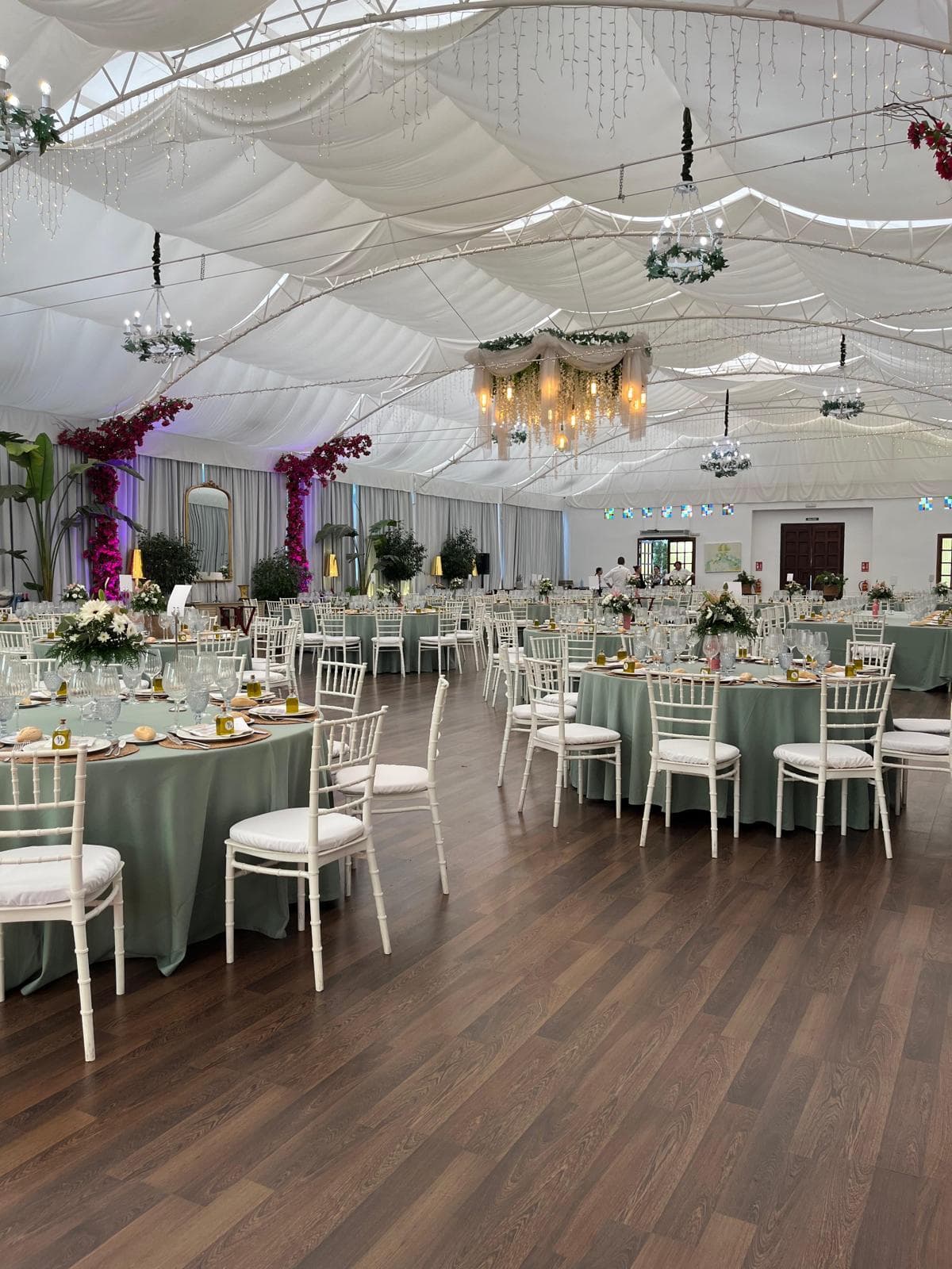 Salón de banquetes con techo de tela drapeada, suelo de madera, mesas redondas con mantel verde salvia y columnas cubiertas de buganvilla