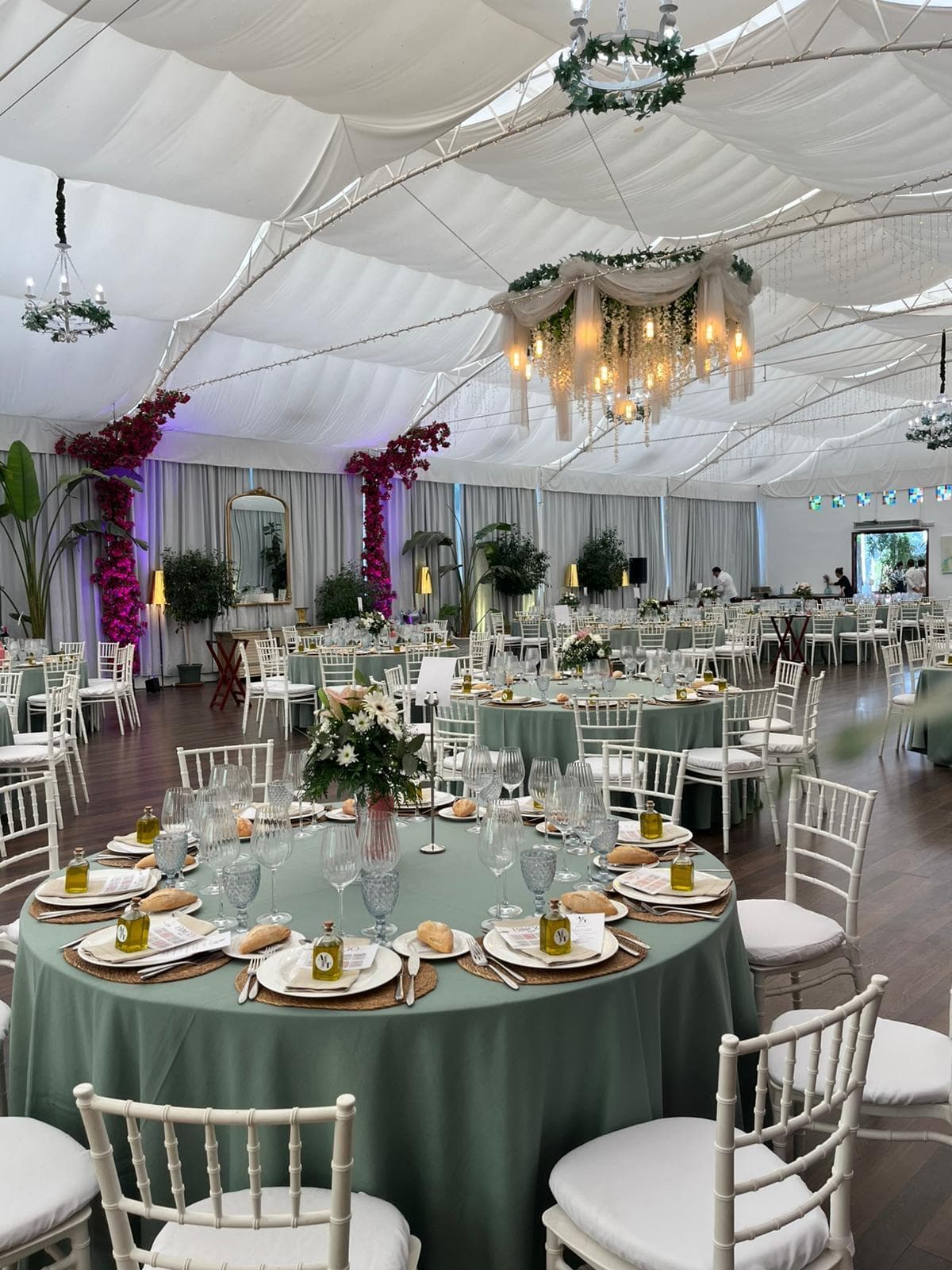 Mesa redonda vestida con mantel verde salvia, vajilla, copas y centro floral, bajo una lámpara de araña de cristal