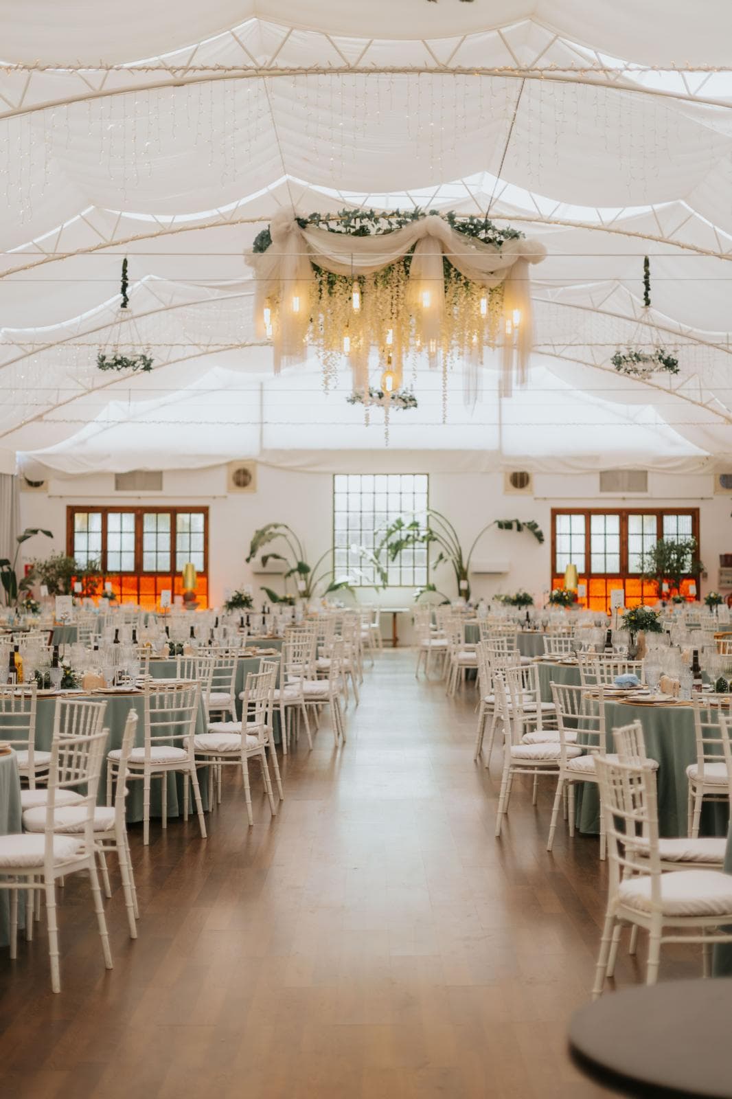 Salón claro con lámpara de flecos, suelo de madera y ventanales al fondo, montado con sillas blancas