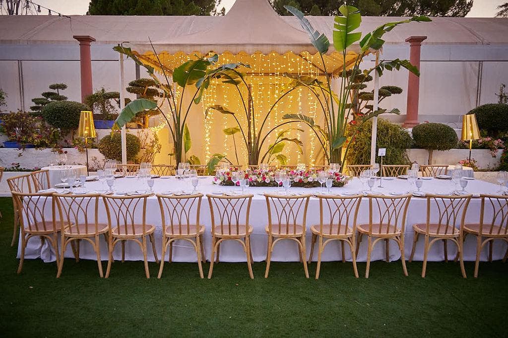 Mesa imperial larga con mantel blanco, sillas de madera y decoración dorada y vegetal en el centro
