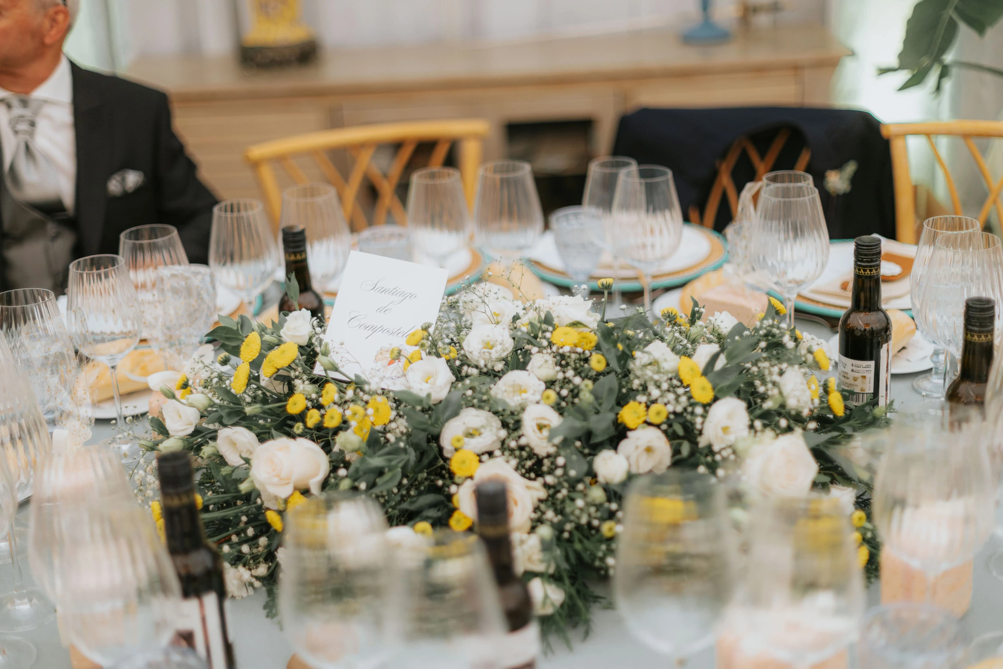 Detalle de mesa presidencial con centro de flores blancas y amarillas, copas y botellas de vino