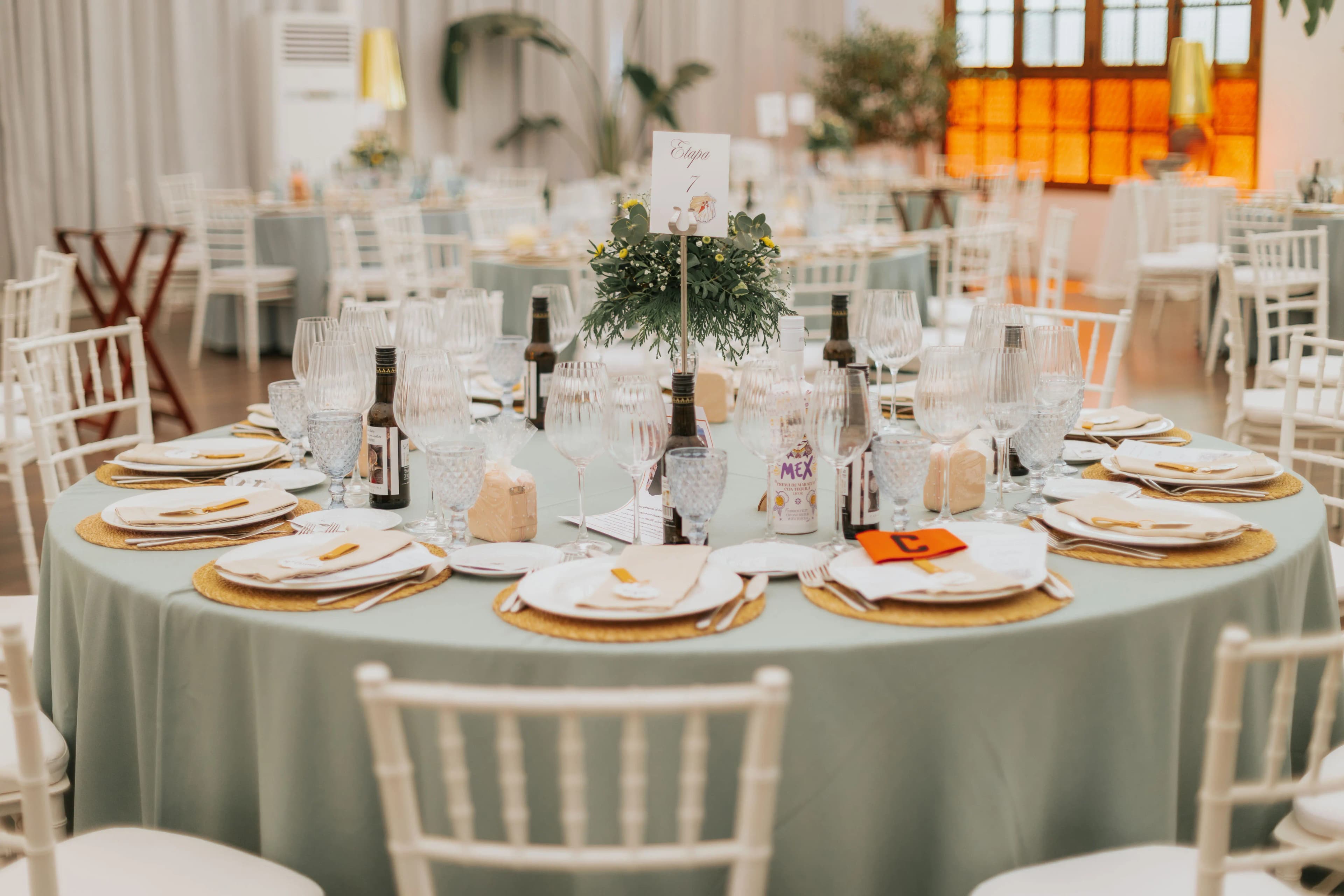 Salón con mesas redondas de mantel verde salvia y sillas blancas, con la luz cálida de los ventanales al fondo