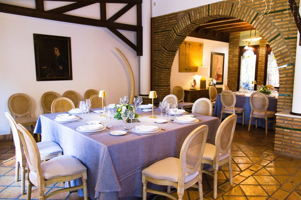 Salón social con techo de tela drapeada, suelo de madera, mesas redondas con mantel verde salvia y columnas cubiertas de buganvilla