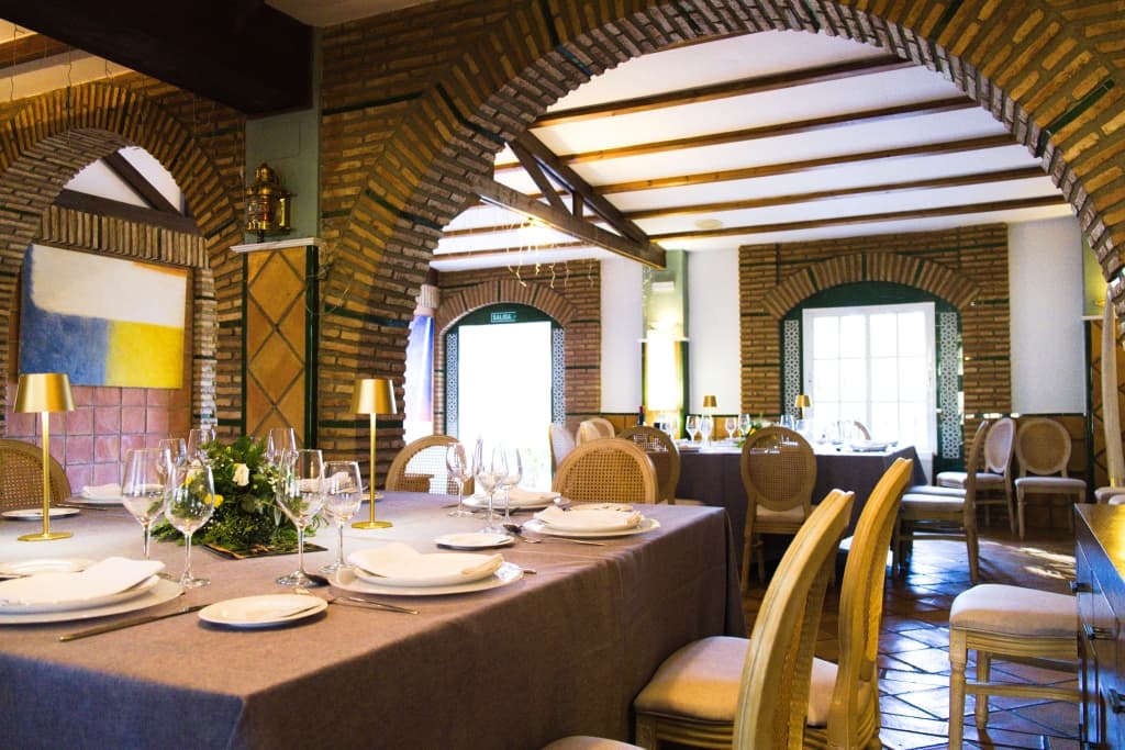 Salón social con techo de tela drapeada, suelo de madera, mesas redondas con mantel verde salvia y columnas cubiertas de buganvilla
