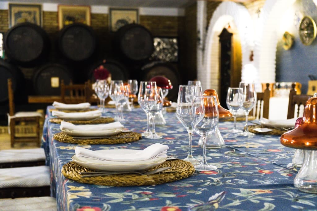 Salón bodeguita con mesa redonda vestida con mantel verde salvia, vajilla, copas y centro floral, bajo una lámpara de araña de cristal