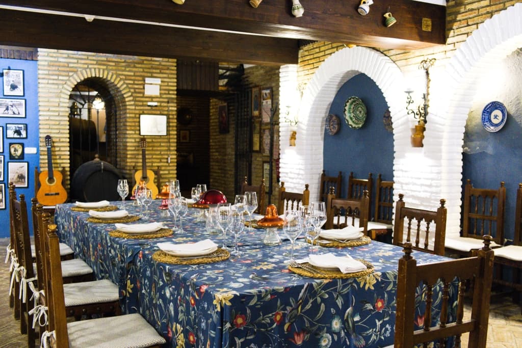 Salón bodeguita con mesa redonda vestida con mantel verde salvia, vajilla, copas y centro floral, bajo una lámpara de araña de cristal