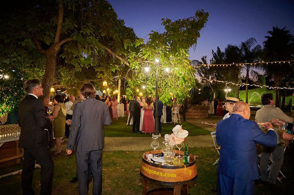 Corte de la tarta nupcial de noche en el jardín, rodeado de invitados bajo guirnaldas de bombillas