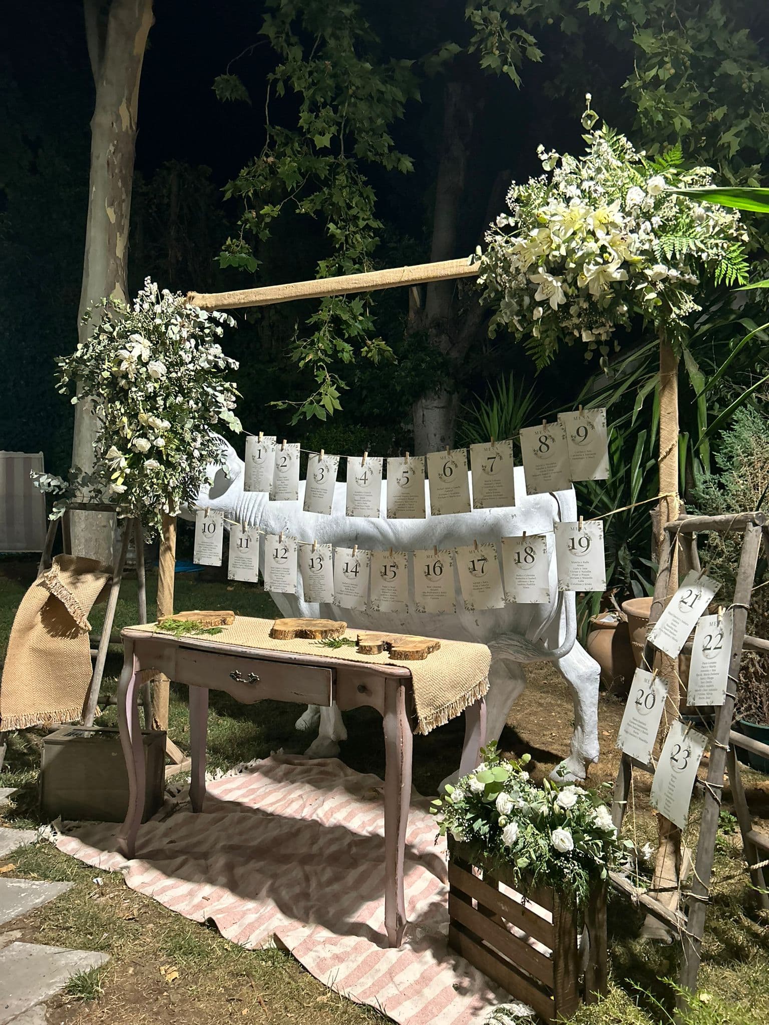 Mesa con el plan de mesas colgado, flores blancas y decoración rústica de madera, iluminada de noche