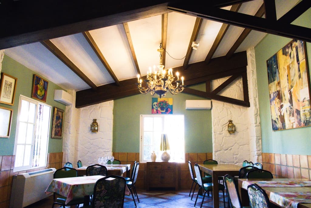 Comedor con techo de vigas de madera, lámpara de araña de forja, cuadros de colores y mesas vestidas