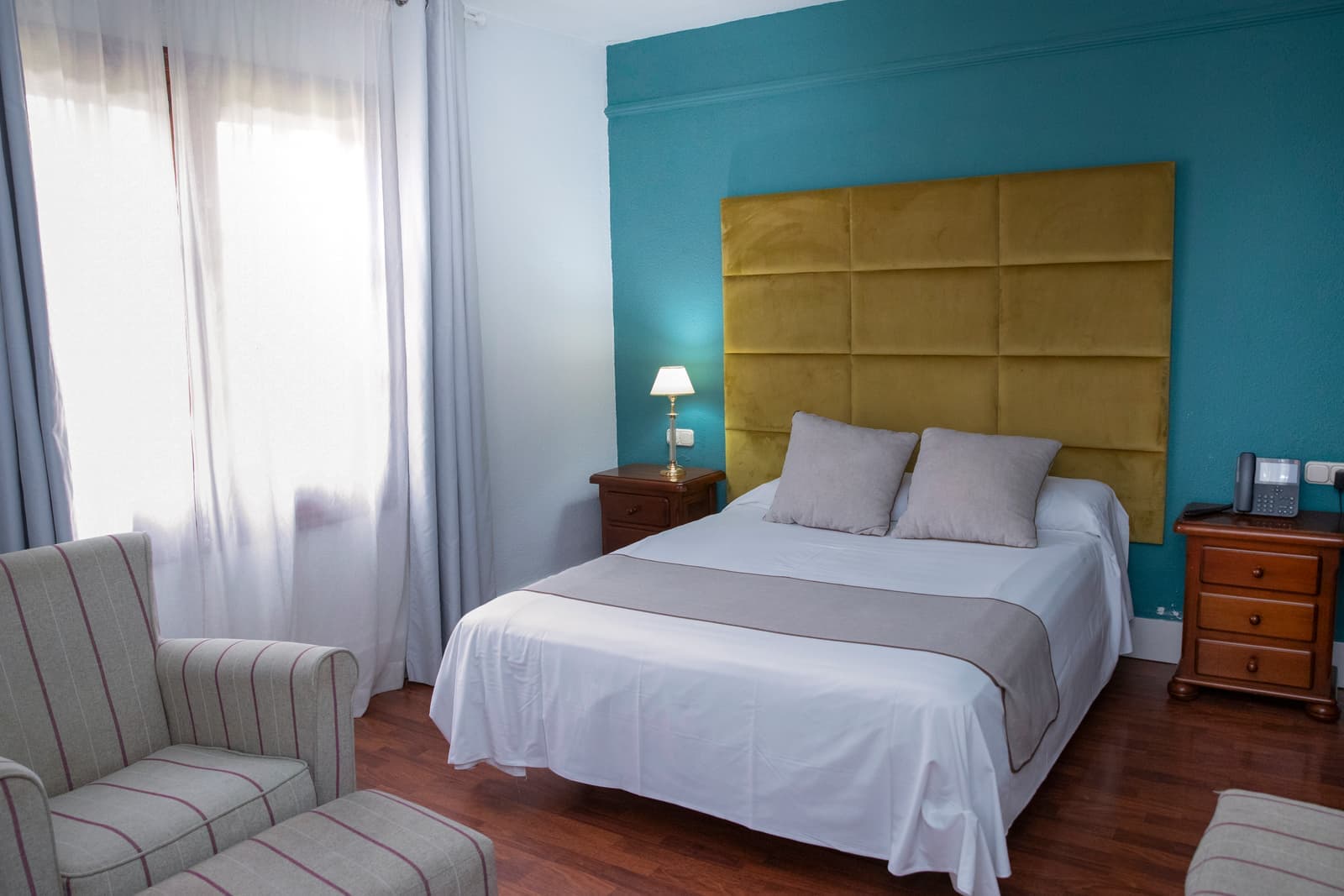 Habitación con pared turquesa, cabecero tapizado en amarillo, cama de matrimonio y butaca junto a la ventana