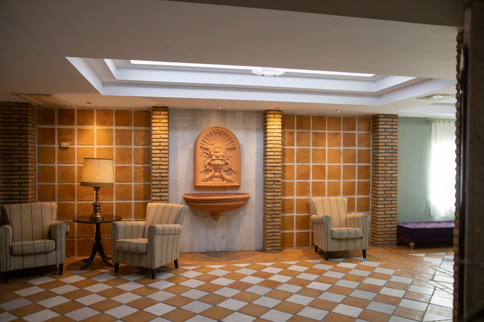 Salón con fuente de piedra sobre pared de ladrillo, butacas tapizadas y suelo de damero