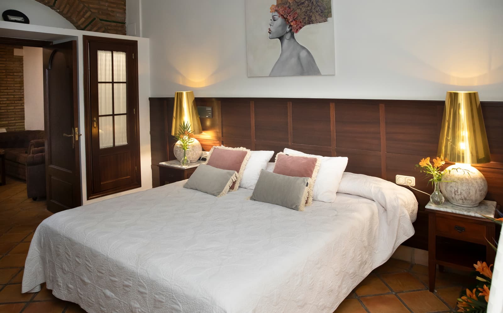 Cama de matrimonio vestida en blanco entre dos lámparas doradas, con cabecero de madera y puerta acristalada al fondo