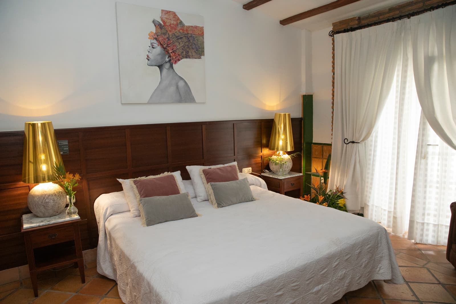 Dormitorio de la suite con cama de matrimonio, lámparas doradas y ventanal con cortinas blancas